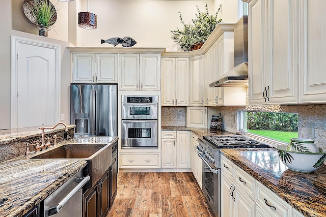 kitchen featuring sink, wall chimney range hood, dark stone countertops, appliances with stainless steel finishes, and light wood-type flooring