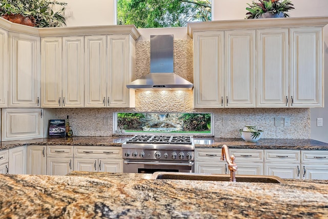 kitchen featuring backsplash, dark stone counters, wall chimney range hood, sink, and stainless steel range