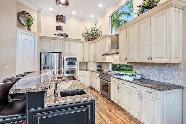 kitchen featuring wall chimney range hood, dark stone countertops, an island with sink, a breakfast bar area, and appliances with stainless steel finishes