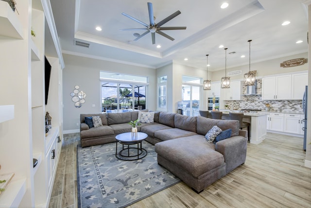 living room with ceiling fan, ornamental molding, a raised ceiling, and light wood-type flooring