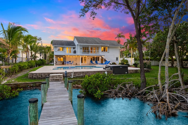 back house at dusk with a swimming pool with hot tub, a balcony, a water view, and a patio