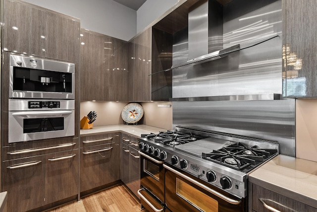 kitchen with dark brown cabinets, stainless steel appliances, light hardwood / wood-style flooring, and wall chimney exhaust hood