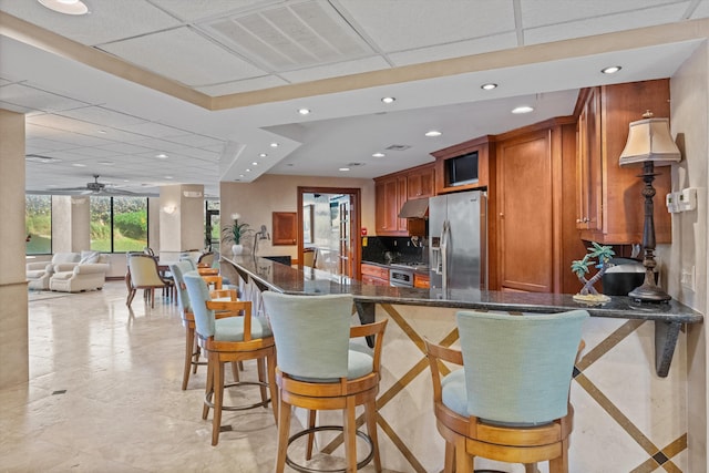 kitchen with ceiling fan, stainless steel fridge with ice dispenser, dark stone countertops, decorative backsplash, and a breakfast bar