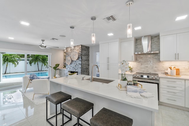 kitchen with wall chimney exhaust hood, ceiling fan, pendant lighting, white cabinetry, and an island with sink