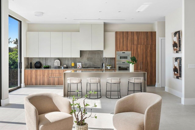 kitchen featuring white cabinetry, stainless steel double oven, a kitchen breakfast bar, backsplash, and a center island with sink