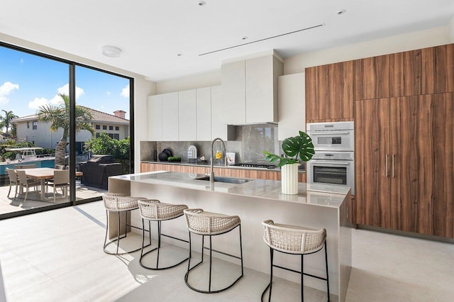 kitchen featuring floor to ceiling windows, a kitchen island with sink, sink, tasteful backsplash, and white cabinetry