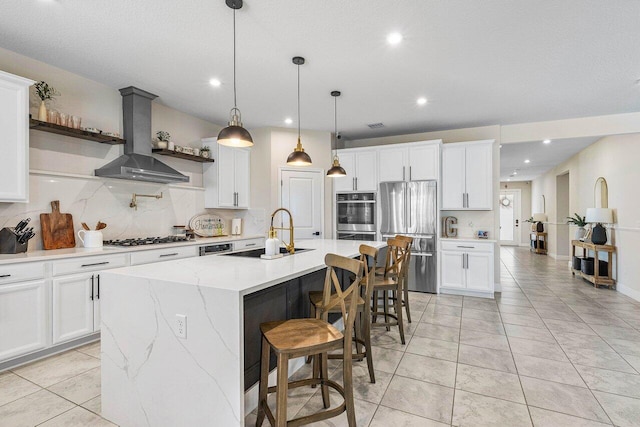 kitchen featuring a center island with sink, white cabinets, wall chimney range hood, and appliances with stainless steel finishes