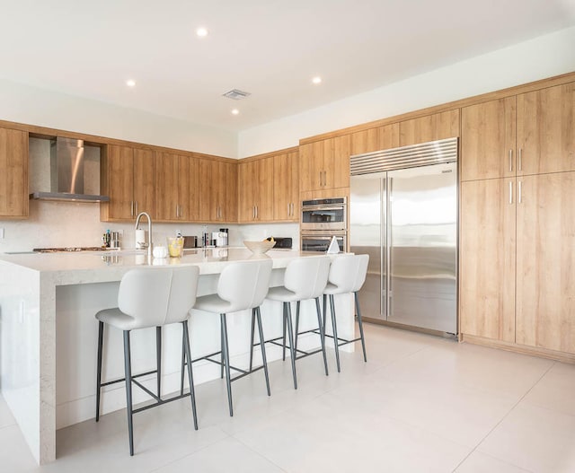 kitchen featuring a kitchen bar, appliances with stainless steel finishes, wall chimney range hood, light tile patterned floors, and an island with sink