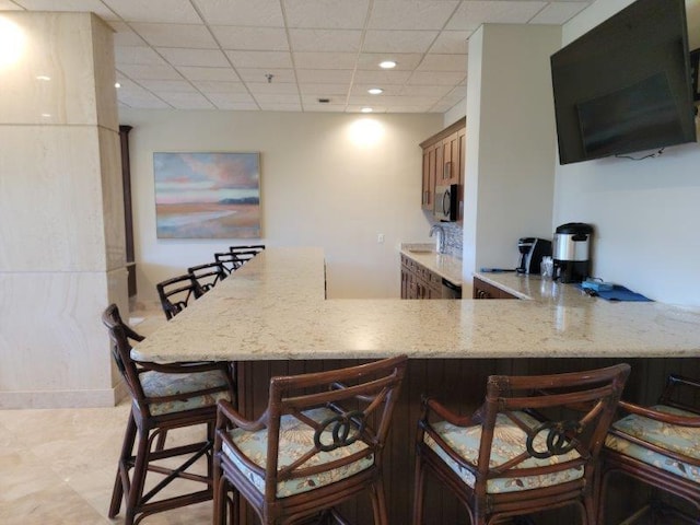 kitchen featuring a drop ceiling, light stone counters, kitchen peninsula, a kitchen bar, and light tile patterned floors