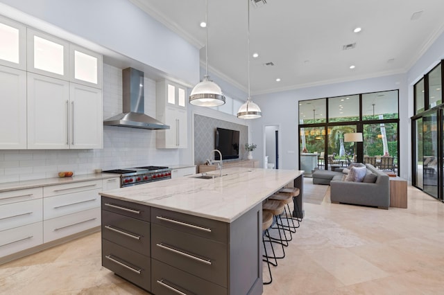 kitchen featuring backsplash, wall chimney range hood, white cabinetry, hanging light fixtures, and an island with sink