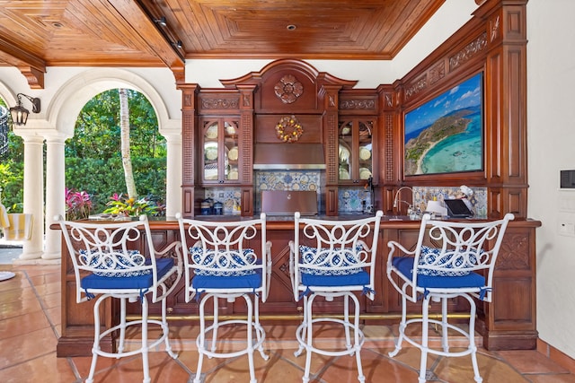 tiled dining space featuring bar area, wooden ceiling, ornamental molding, and ornate columns