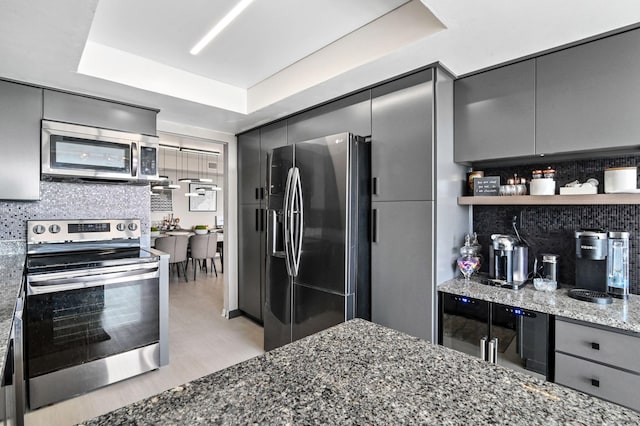 kitchen with appliances with stainless steel finishes, backsplash, dark stone counters, a tray ceiling, and gray cabinets