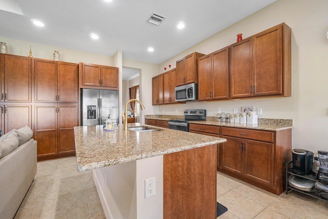 kitchen featuring light stone countertops, a textured ceiling, stainless steel appliances, sink, and an island with sink