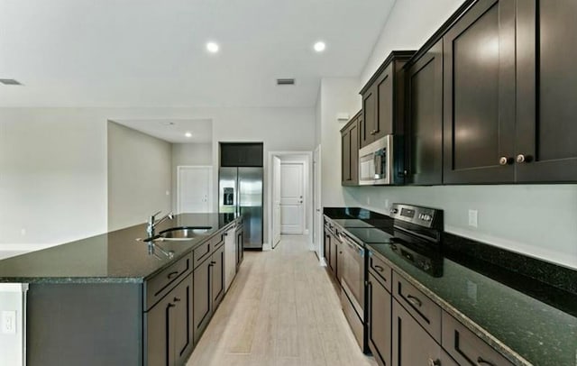 kitchen featuring light wood-type flooring, stainless steel appliances, a kitchen island with sink, sink, and dark stone countertops