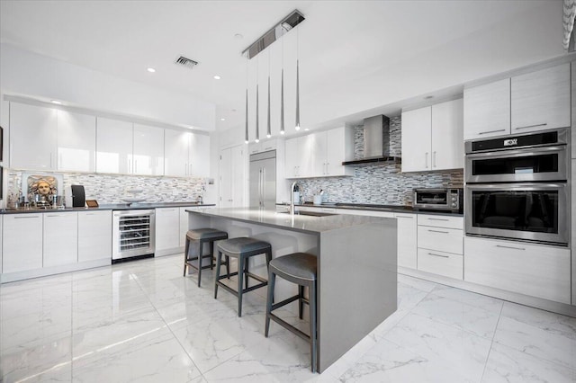 kitchen with wall chimney range hood, hanging light fixtures, an island with sink, stainless steel appliances, and beverage cooler