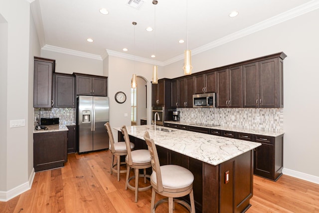 kitchen featuring light wood-type flooring, ornamental molding, stainless steel appliances, decorative light fixtures, and a center island with sink