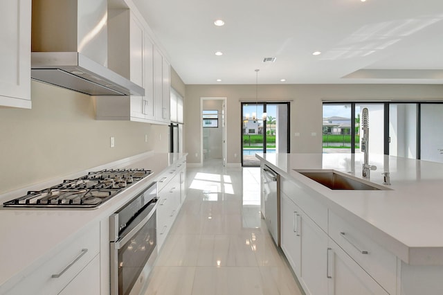 kitchen with white cabinets, wall chimney range hood, sink, hanging light fixtures, and stainless steel appliances