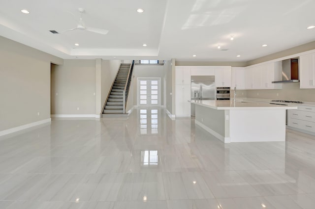 kitchen featuring a large island with sink, wall chimney exhaust hood, ceiling fan, appliances with stainless steel finishes, and white cabinetry