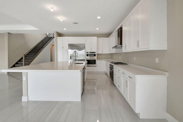 kitchen with white cabinets, wall chimney range hood, sink, an island with sink, and appliances with stainless steel finishes