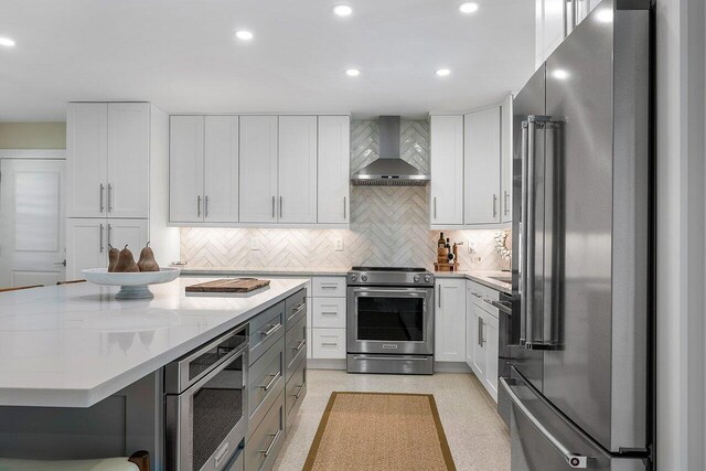 kitchen featuring backsplash, wall chimney range hood, light stone countertops, appliances with stainless steel finishes, and white cabinetry