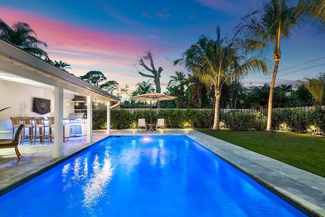 pool at dusk featuring area for grilling, exterior kitchen, a bar, a patio area, and a yard