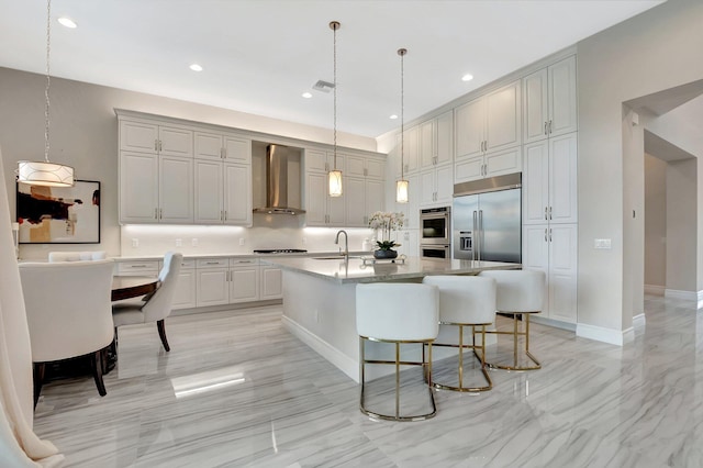 kitchen with built in fridge, gray cabinetry, wall chimney range hood, a center island with sink, and pendant lighting