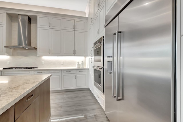 kitchen with stainless steel appliances, backsplash, white cabinetry, wall chimney range hood, and light stone countertops