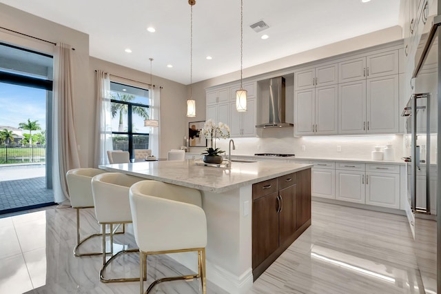 kitchen featuring visible vents, an island with sink, wall chimney exhaust hood, decorative light fixtures, and light stone countertops