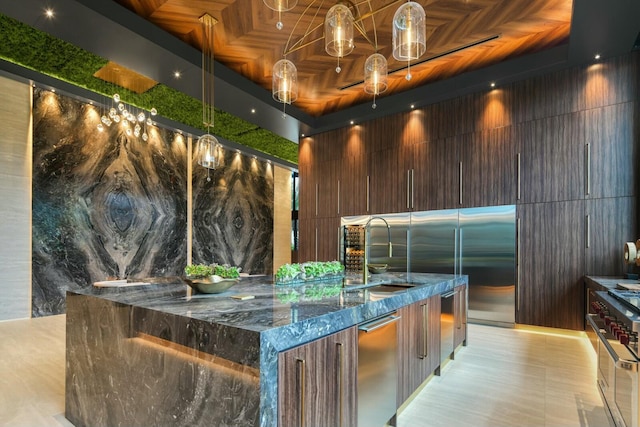 kitchen featuring high end appliances, a large island with sink, sink, dark stone countertops, and dark brown cabinetry