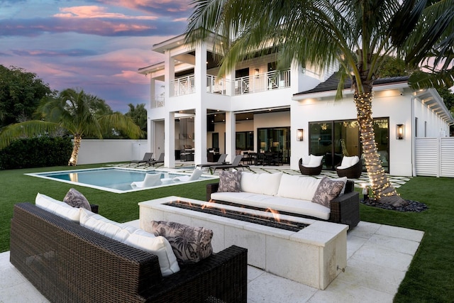 back house at dusk with a lawn, a balcony, an outdoor living space with a fire pit, a fenced in pool, and a patio area