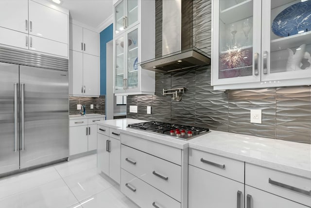 kitchen with wall chimney range hood, white cabinetry, backsplash, stainless steel appliances, and light stone counters