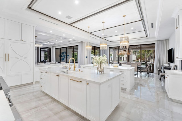 kitchen with light stone counters, sink, white cabinets, hanging light fixtures, and an island with sink