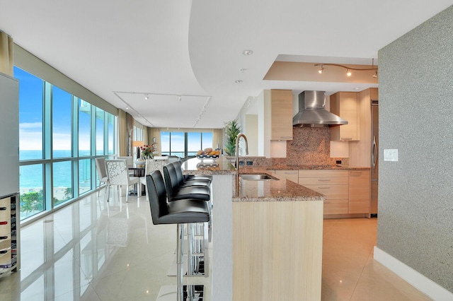 kitchen with light stone countertops, light brown cabinetry, wall chimney exhaust hood, sink, and a water view