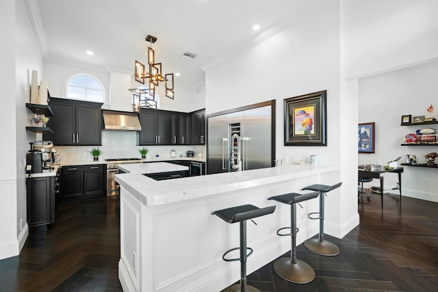 kitchen with kitchen peninsula, dark parquet flooring, wall chimney exhaust hood, a breakfast bar, and decorative light fixtures