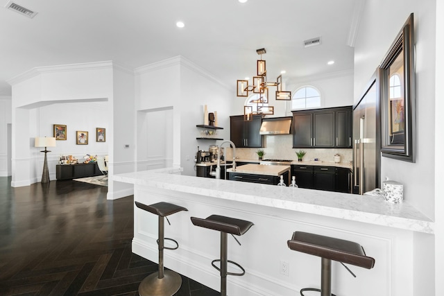 kitchen with dark parquet flooring, wall chimney range hood, crown molding, decorative light fixtures, and a breakfast bar area