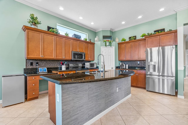 kitchen featuring decorative backsplash, appliances with stainless steel finishes, dark stone counters, a kitchen island with sink, and light tile patterned floors