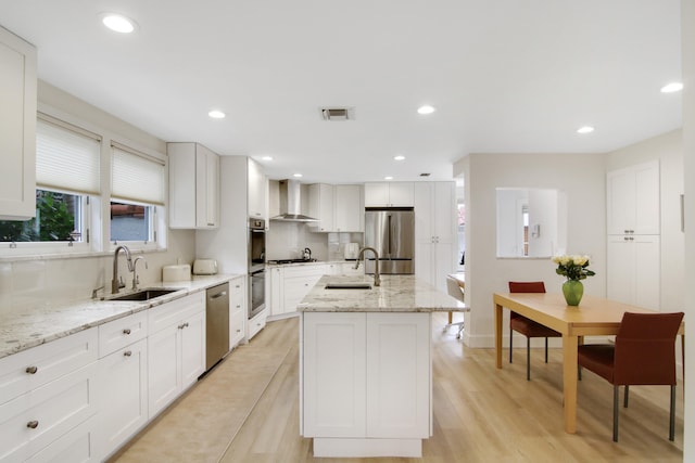 kitchen featuring a center island with sink, wall chimney range hood, sink, light wood-type flooring, and appliances with stainless steel finishes