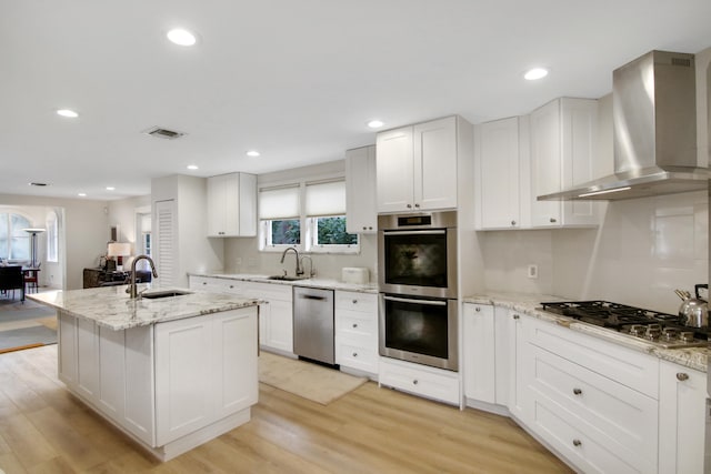 kitchen featuring light hardwood / wood-style flooring, an island with sink, wall chimney range hood, and appliances with stainless steel finishes