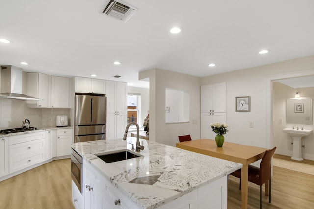 kitchen with stainless steel appliances, a kitchen island with sink, sink, wall chimney range hood, and light hardwood / wood-style flooring