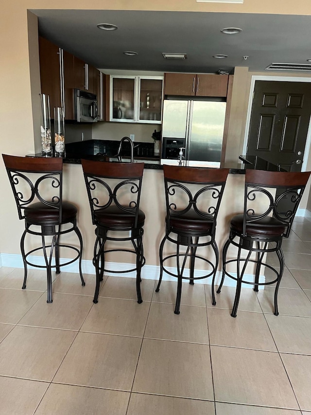 kitchen featuring kitchen peninsula, a kitchen bar, dark brown cabinetry, stainless steel appliances, and light tile patterned floors