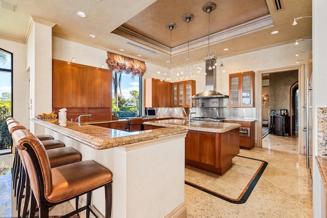 kitchen with kitchen peninsula, wall chimney exhaust hood, a tray ceiling, crown molding, and decorative light fixtures