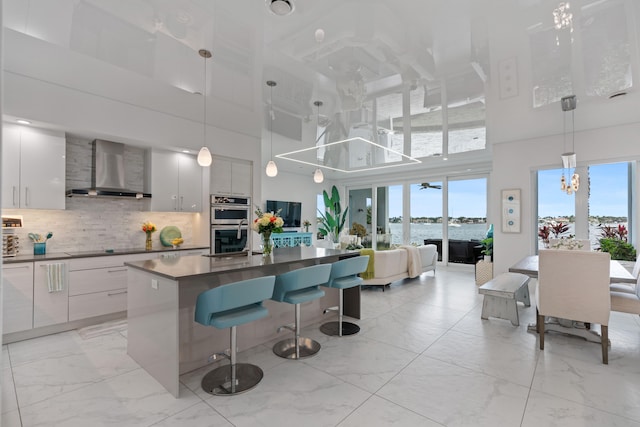 kitchen featuring white cabinets, a towering ceiling, wall chimney exhaust hood, and hanging light fixtures