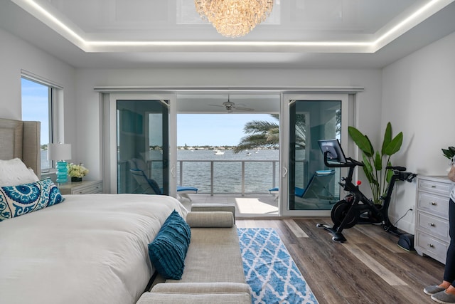 bedroom with access to exterior, dark hardwood / wood-style flooring, a tray ceiling, a water view, and a chandelier