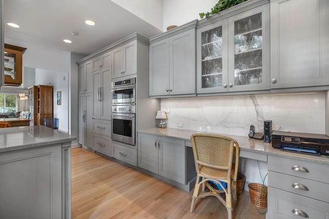 kitchen featuring light stone countertops, stainless steel double oven, gray cabinets, built in desk, and light wood-type flooring