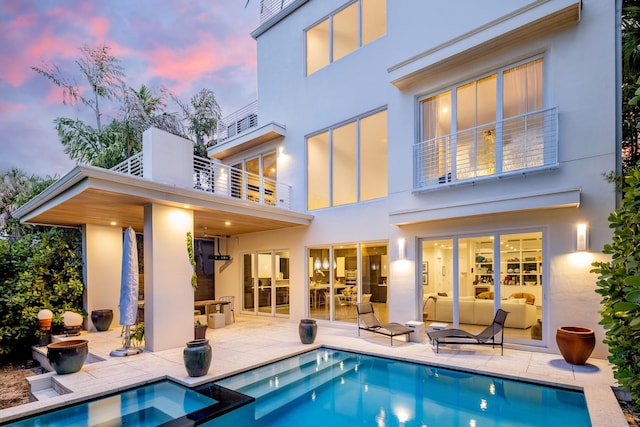 back house at dusk featuring a swimming pool with hot tub, a patio area, and a balcony