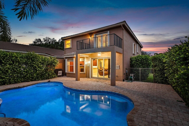 back house at dusk with ceiling fan, a balcony, a fenced in pool, and a patio