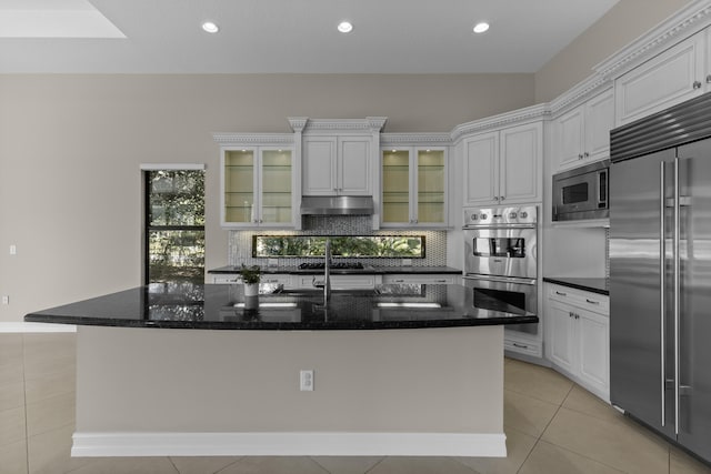 kitchen with light tile patterned floors, built in appliances, an island with sink, and white cabinets