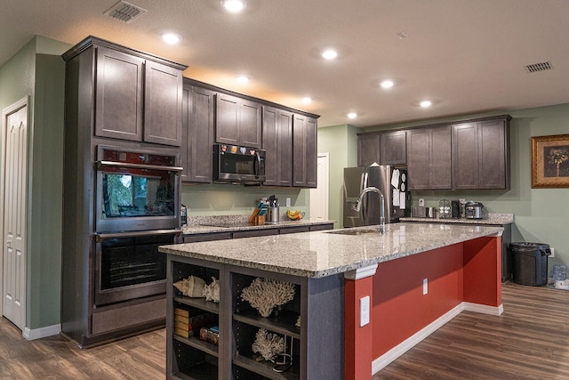 kitchen featuring appliances with stainless steel finishes, light stone counters, dark brown cabinetry, dark wood-type flooring, and an island with sink