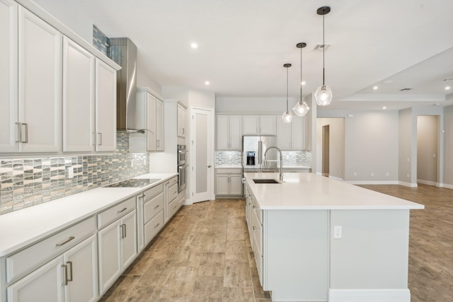 kitchen featuring wall chimney range hood, sink, a large island with sink, white cabinets, and hanging light fixtures