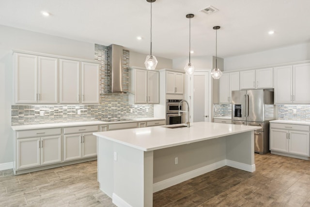 kitchen featuring white cabinetry, hanging light fixtures, wall chimney range hood, a center island with sink, and appliances with stainless steel finishes